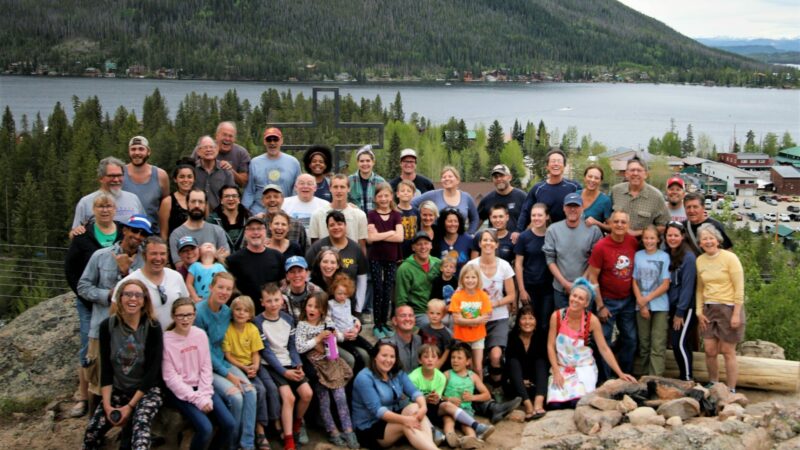 a few dozen people pose for a group photo on the top of a cliff with a lake and mountain in the background